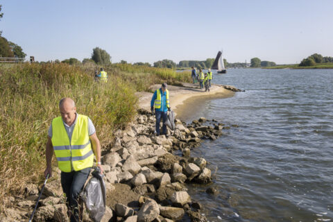 Vrijwilligers in gele hesjes ruimen afval op langs de rivier. Zeilboot op de achtergrond.