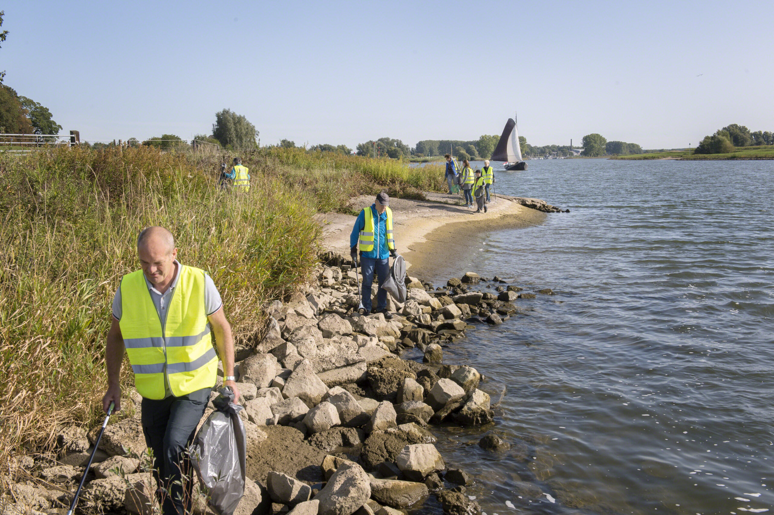Vrijwilligers in gele hesjes ruimen afval op langs de rivier. Zeilboot op de achtergrond.