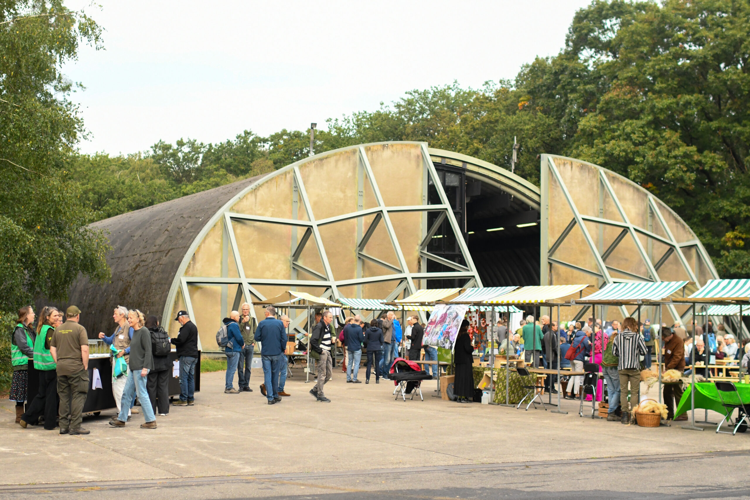Buitenevenement bij een moderne hangar, mensen bij kraampjes, bomen op de achtergrond.