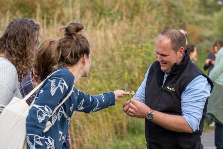 Een man toont mensen een bloem tijdens een natuurwandeling in een grasrijk gebied.