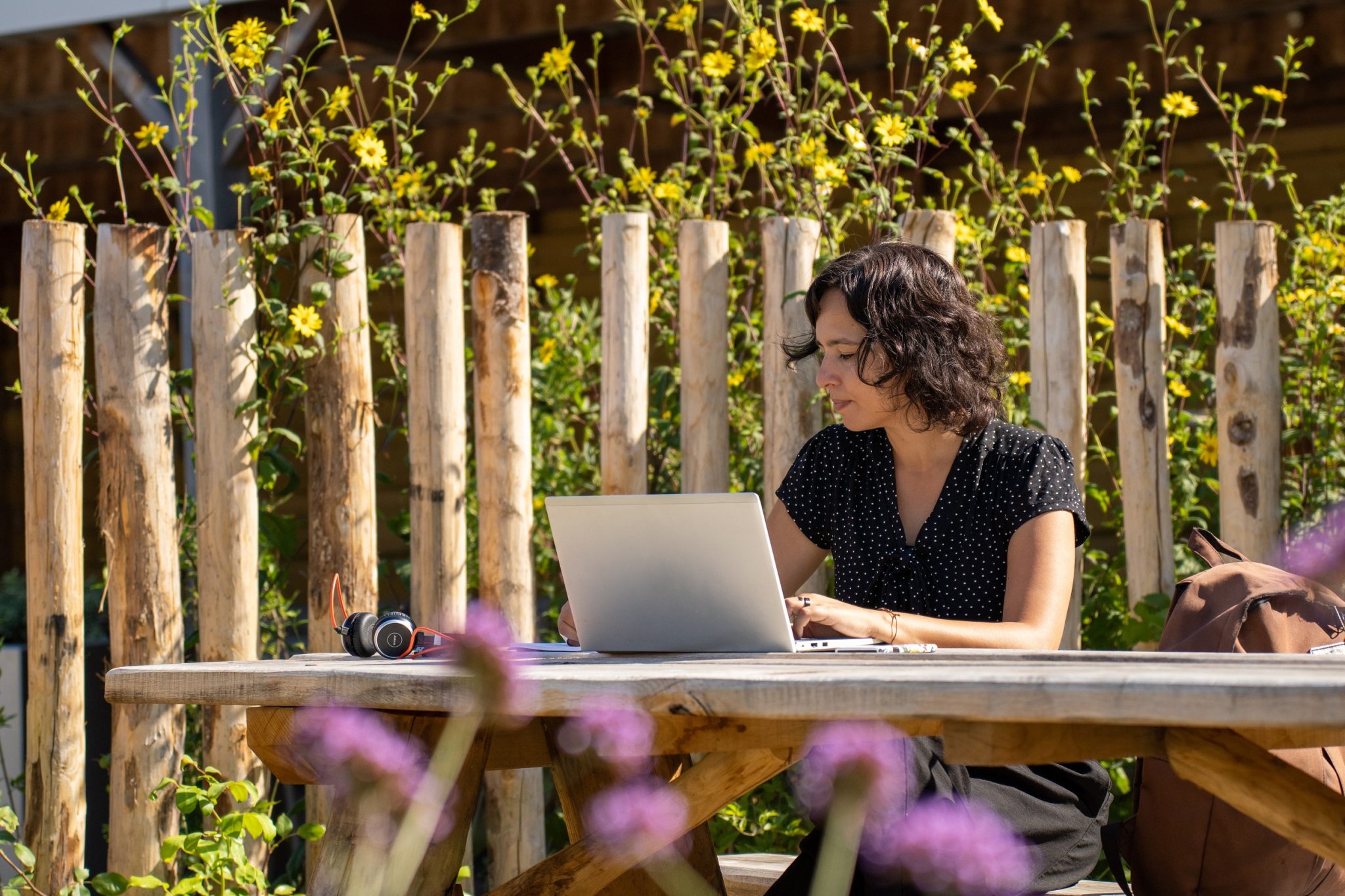 Vrouw werkt op laptop buiten aan houten tafel met bloemen en houten hek op achtergrond.