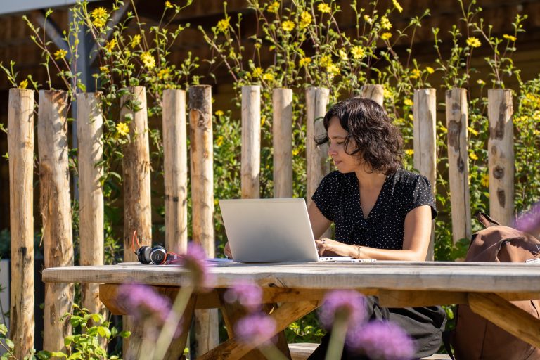 Vrouw werkt op laptop buiten aan houten tafel met bloemen en houten hek op achtergrond.