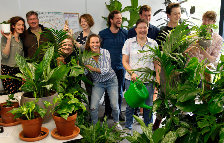 Groep mensen met kamerplanten in een kantoorruimte, vrolijk poserend met groen rondom.
