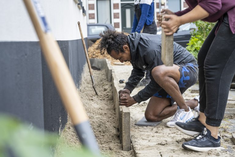 Twee mensen leggen samen stenen in een zandbed naast een muur.