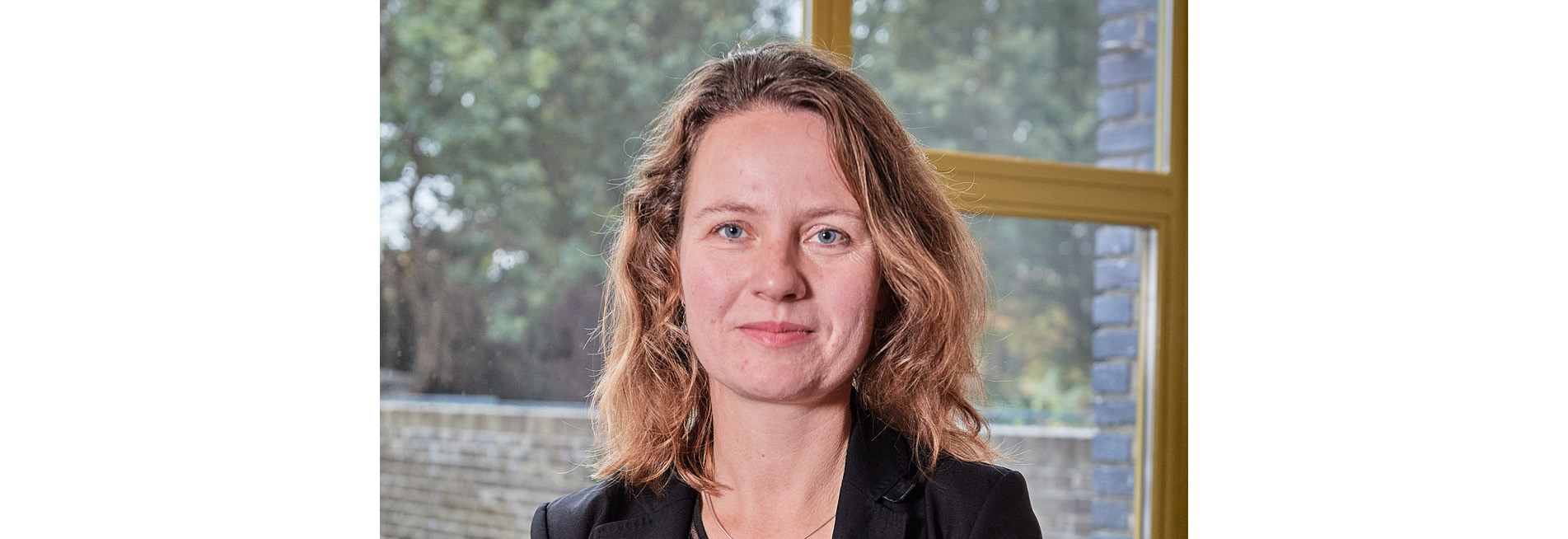 Vrouw met halflang haar in een zwarte blazer voor een raam met groene achtergrond.