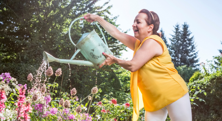 Vrouw in gele top geeft bloemen water in zonnige tuin.