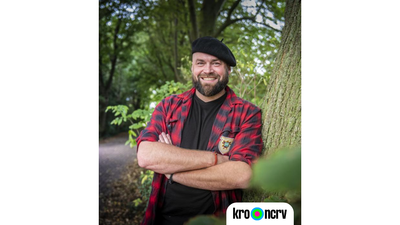 Man met baret, glimlachend, rood geruit overhemd, leunend tegen boom in bos; KRO-NCRV logo.