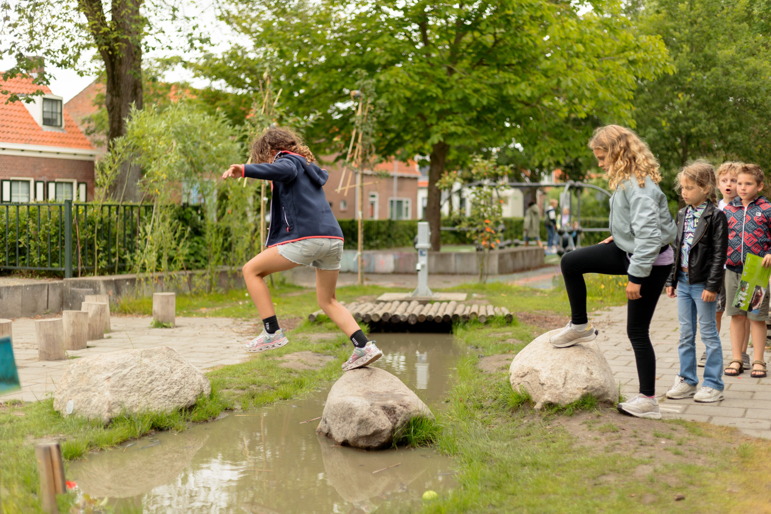 Kinderen spelen en springen over stenen boven een modderige waterplas in een speeltuin.