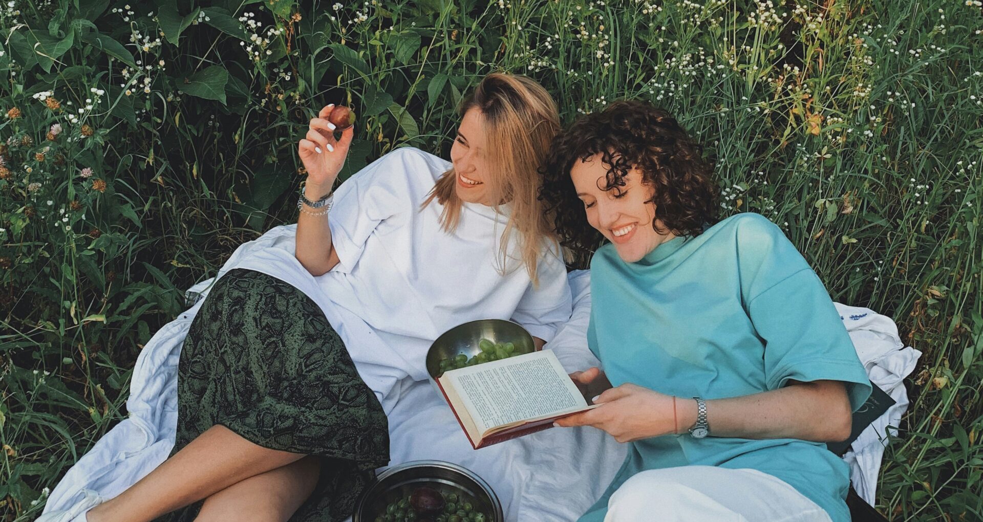 Twee vrouwen ontspannen met een boek en fruit, liggend in een grasveld vol bloemen.
