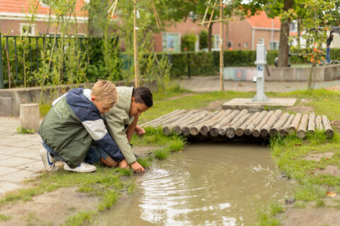 Twee kinderen spelen bij een modderpoel naast een houten brug in een groene tuin.