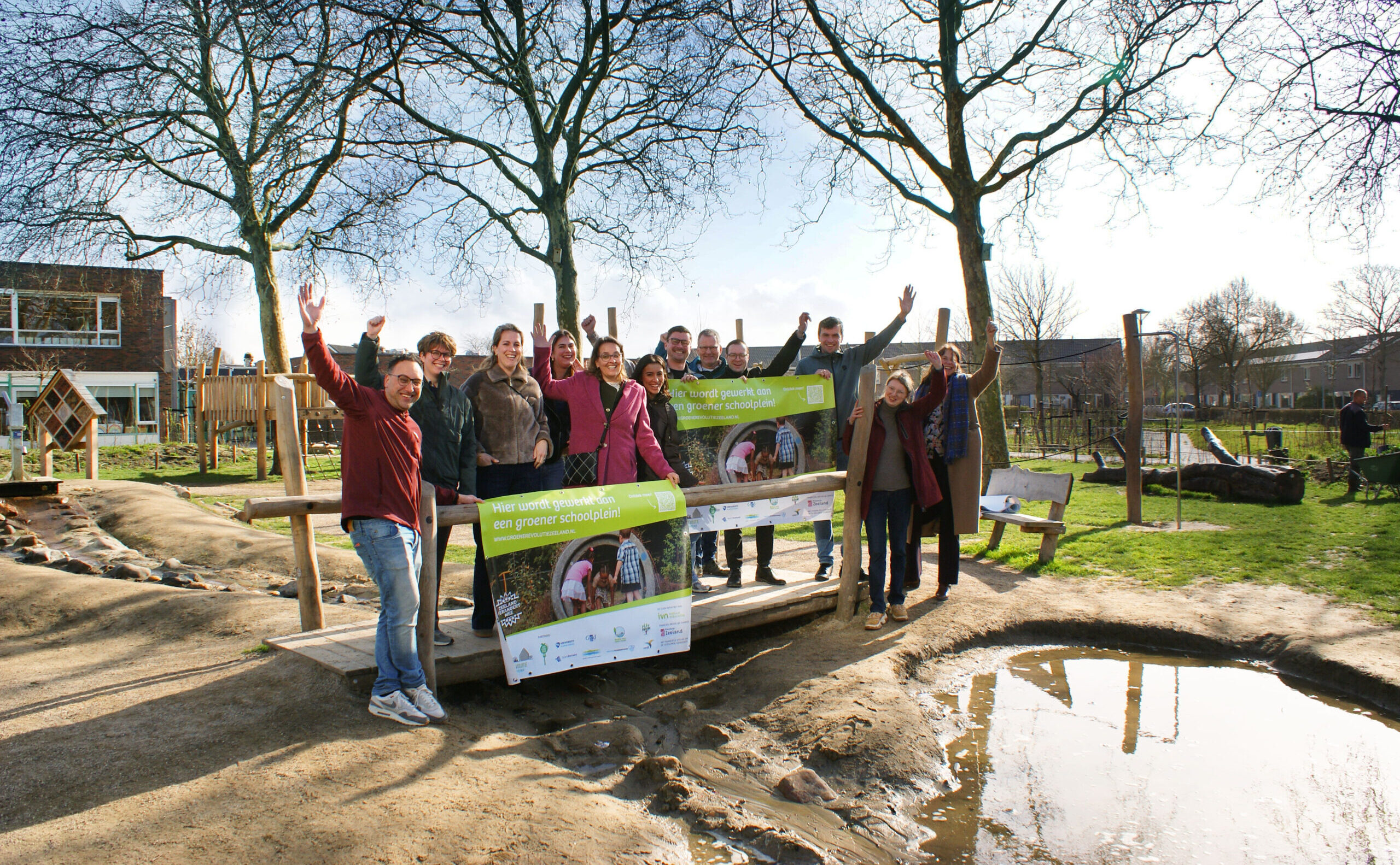 Groep mensen poseert met een spandoek op een groen schoolplein, omringd door bomen.