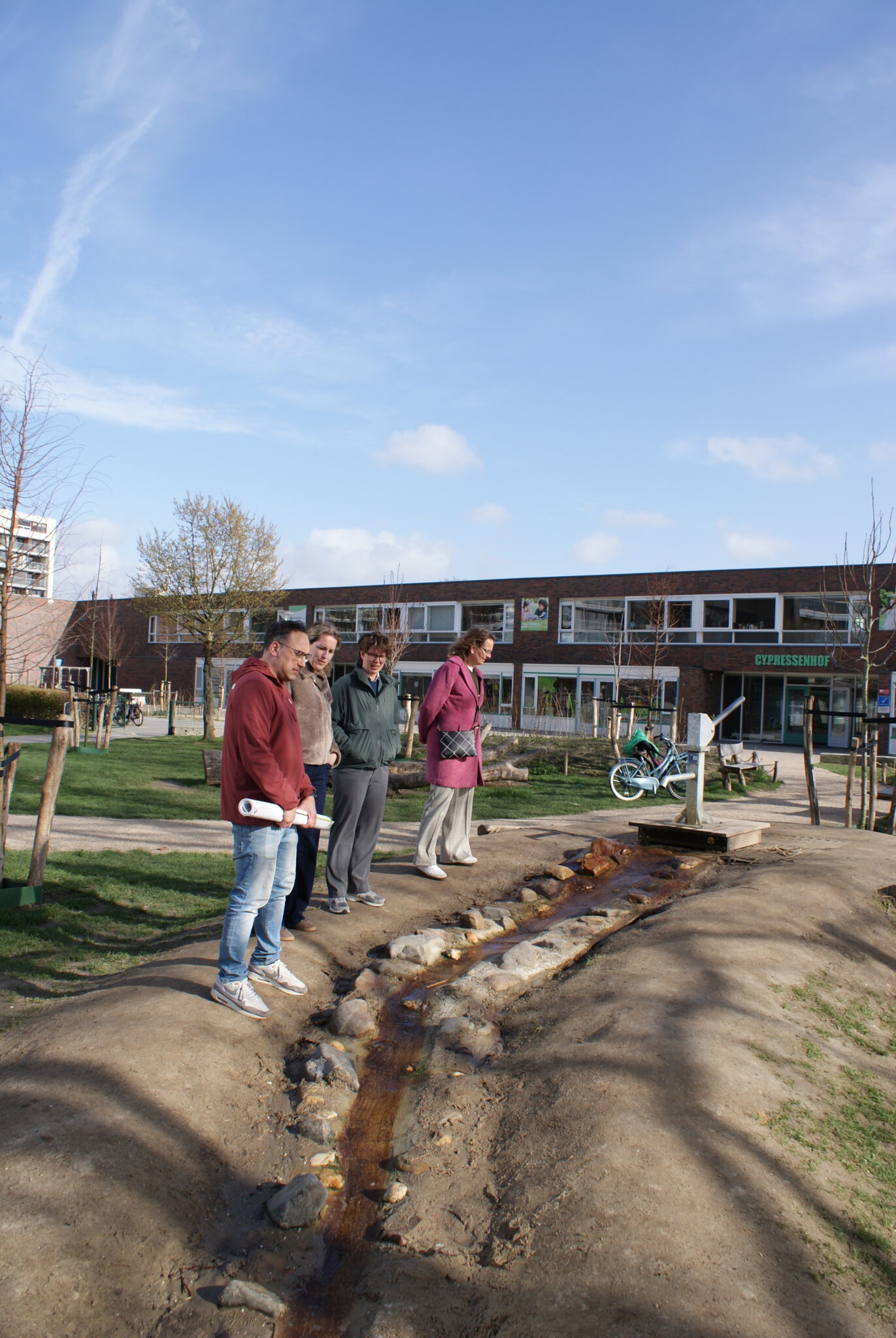 Vier mensen bekijken een kleine waterstroom in een groene buurt, met een school op de achtergrond.