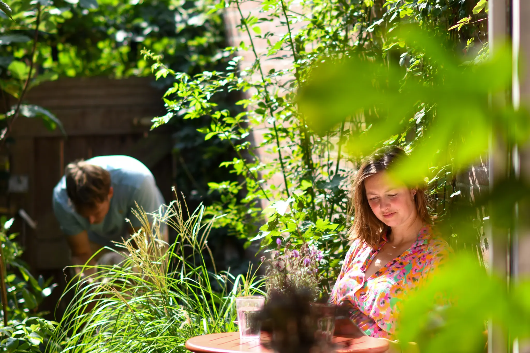 Vrouw leest aan tafel in groene tuin, persoon op achtergrond tuiniert.