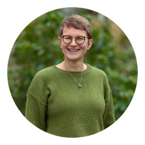 Vrouw met korte bruine haren en bril, lacht in groene trui voor groene achtergrond.
