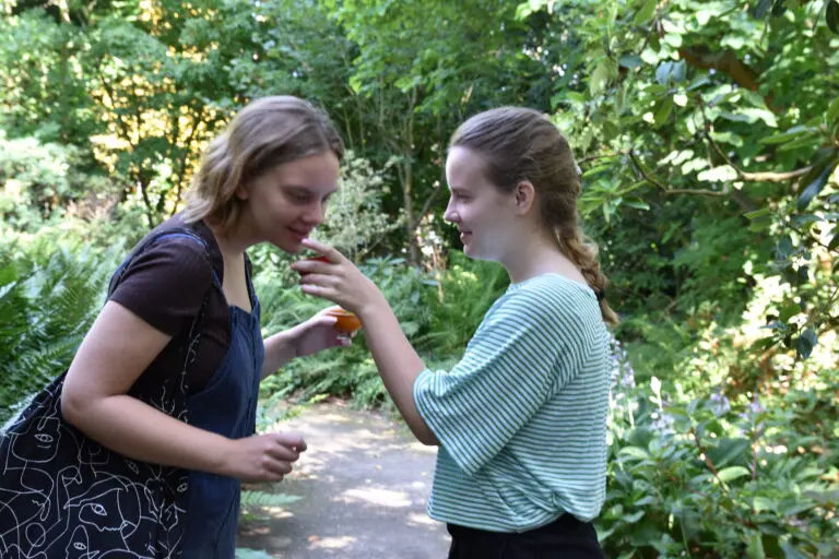 Twee jonge vrouwen in een groene tuin, één biedt de ander iets aan in een klein potje.
