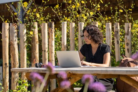 Vrouw werkt buiten aan een laptop op een houten tafel, omgeven door bloemen en een houten hek.