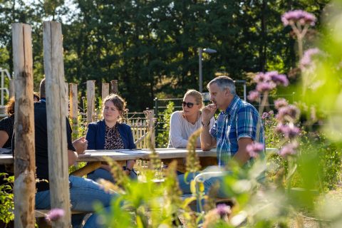 Mensen vergaderen aan een houten tafel in een groene, zonnige tuin omringd door bloemen.