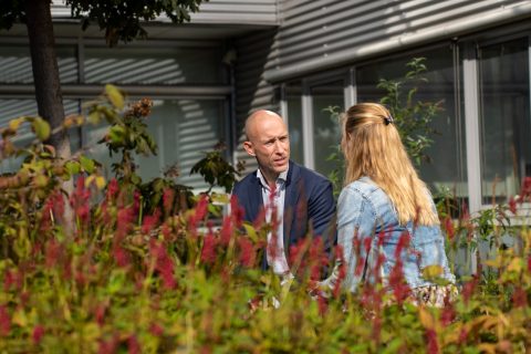 Twee personen in gesprek in een tuin, omgeven door bloemen en een gebouw op de achtergrond.