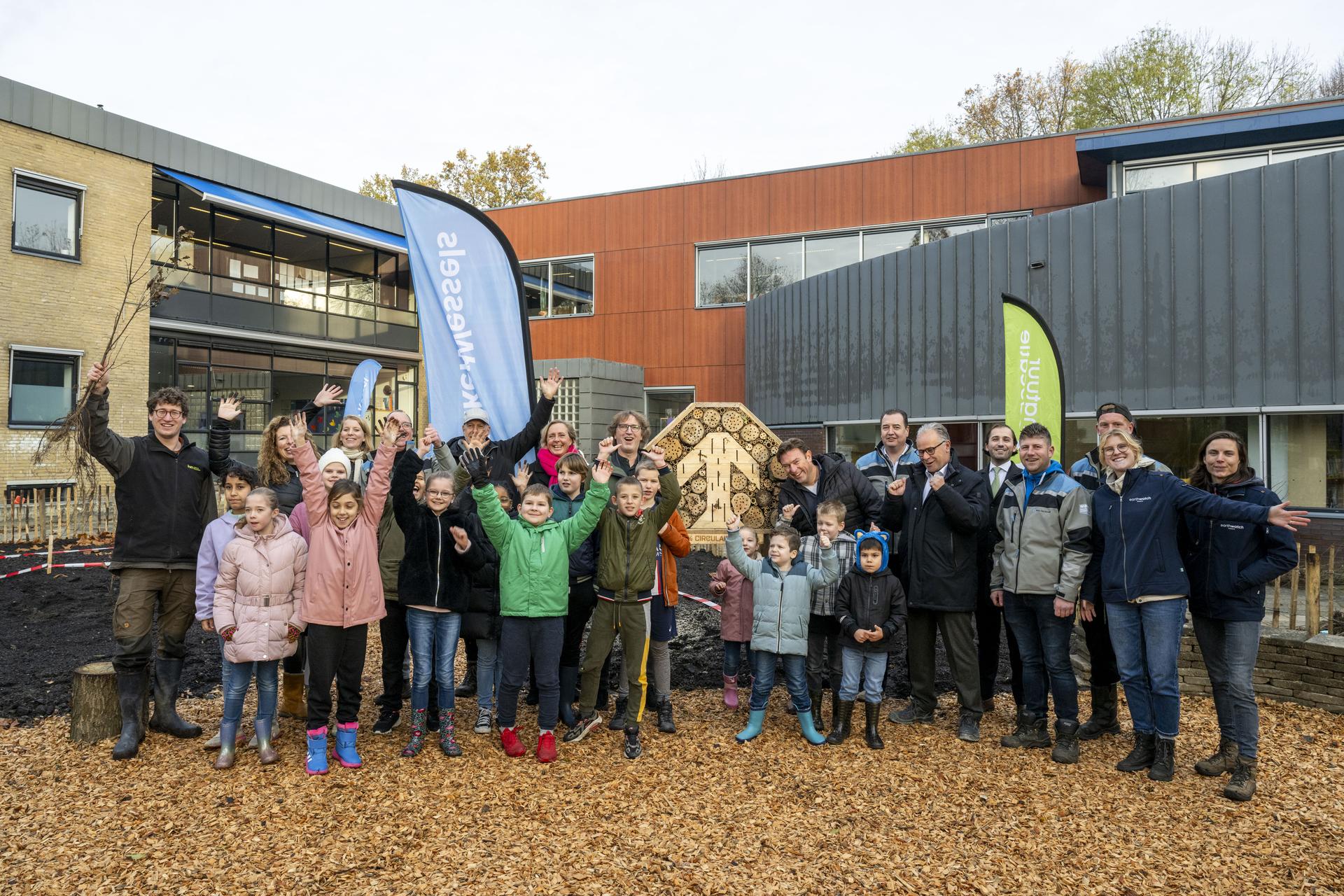 Groep mensen, waaronder kinderen, viert buiten voor een schoolgebouw met banners en een bijenhotel.