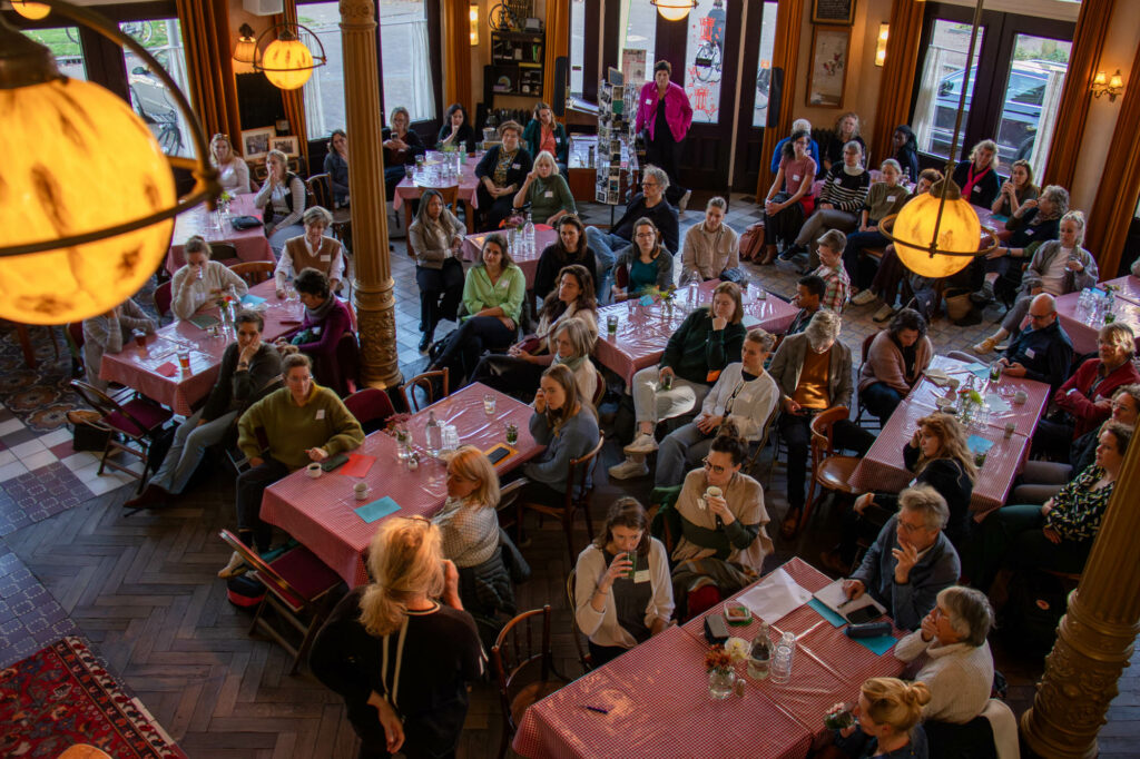 Grote groep mensen luistert aandachtig in een café-achtige ruimte met rood geruite tafels en warme verlichting.