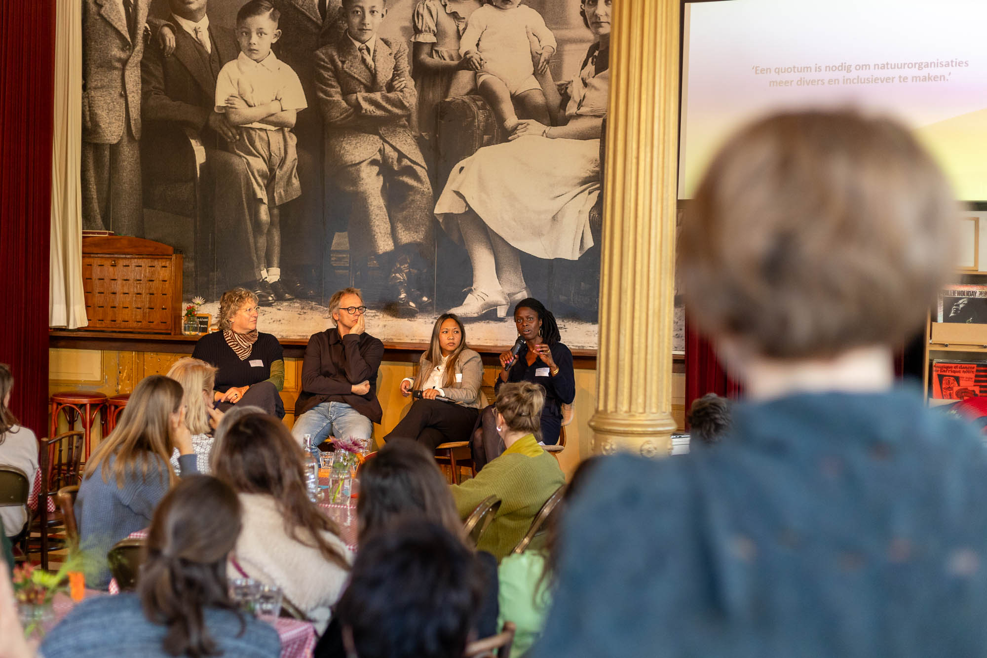 Paneldiscussie met vier sprekers, publiek op de voorgrond, grote familiefoto op de achtergrond.