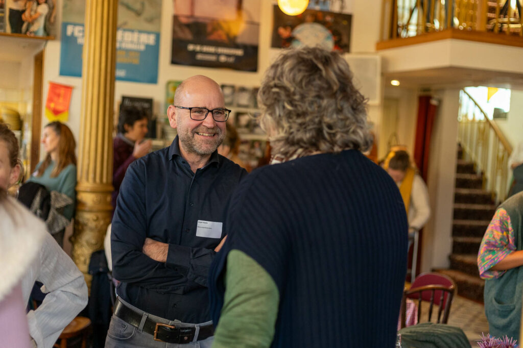 Persoon in gesprek op een drukke bijeenkomst, met posters op de achtergrond.