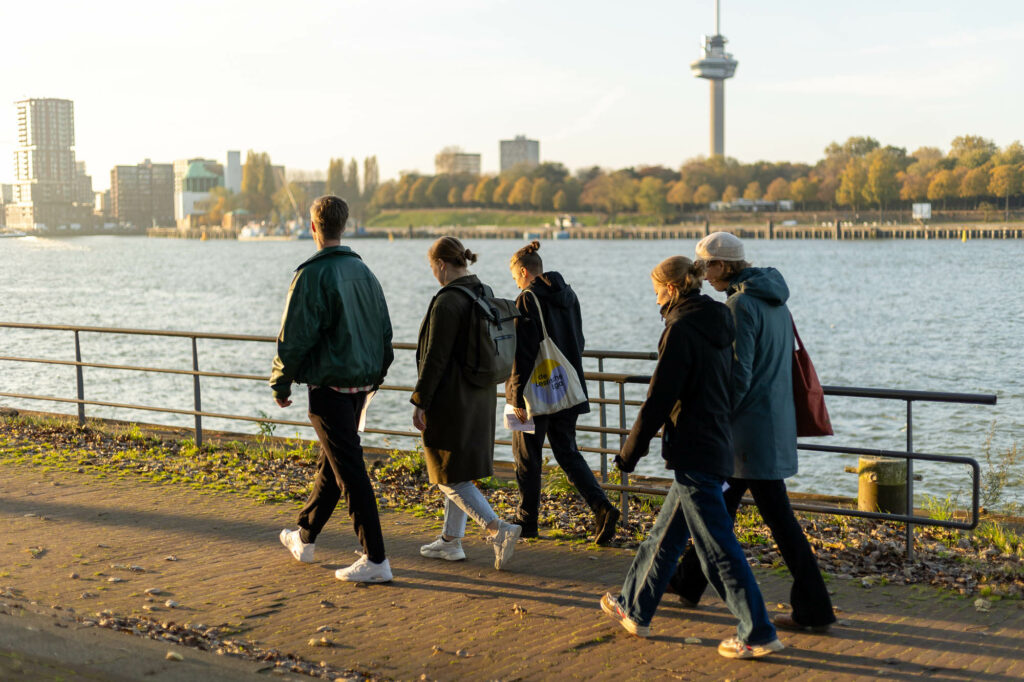 Groep mensen wandelt langs de rivier met een stadsgezicht en toren op de achtergrond.