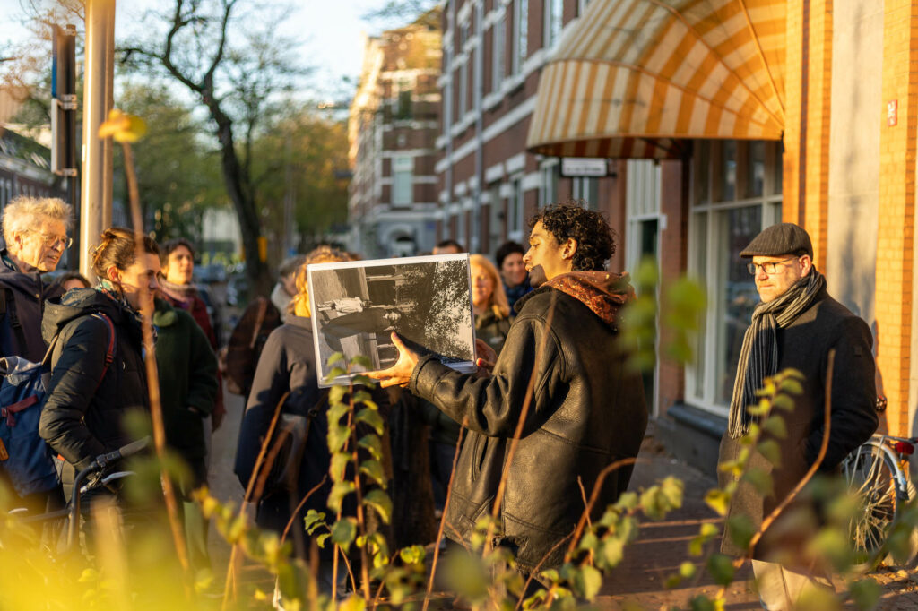 Groep mensen kijkt naar een foto, gepresenteerd door een man op een zonnige straat.