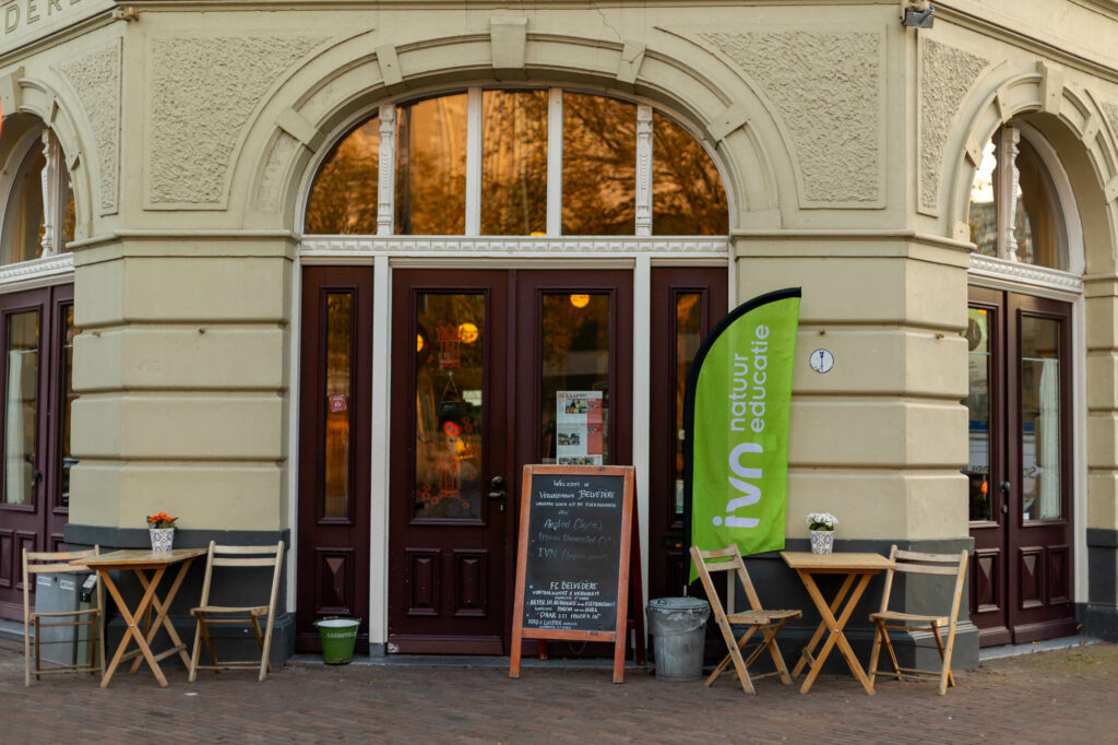 Gevel van een café met houten tafels, groene IVN-vlag en een krijtbord bij de ingang.
