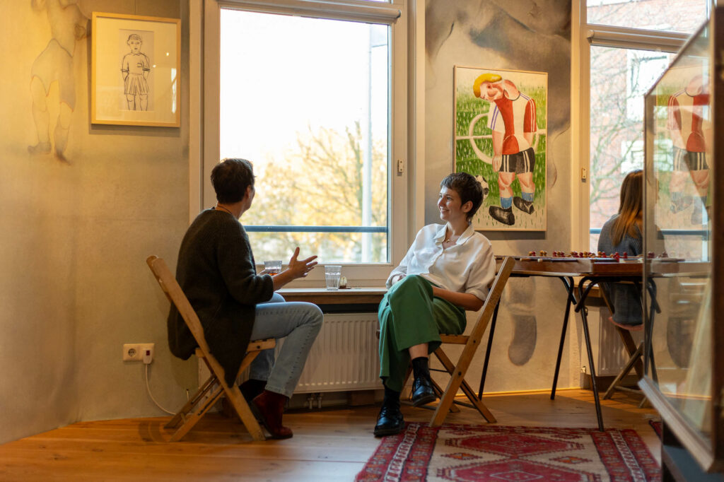 Twee personen zitten op houten stoelen in een kunstgalerij en praten bij het raam.