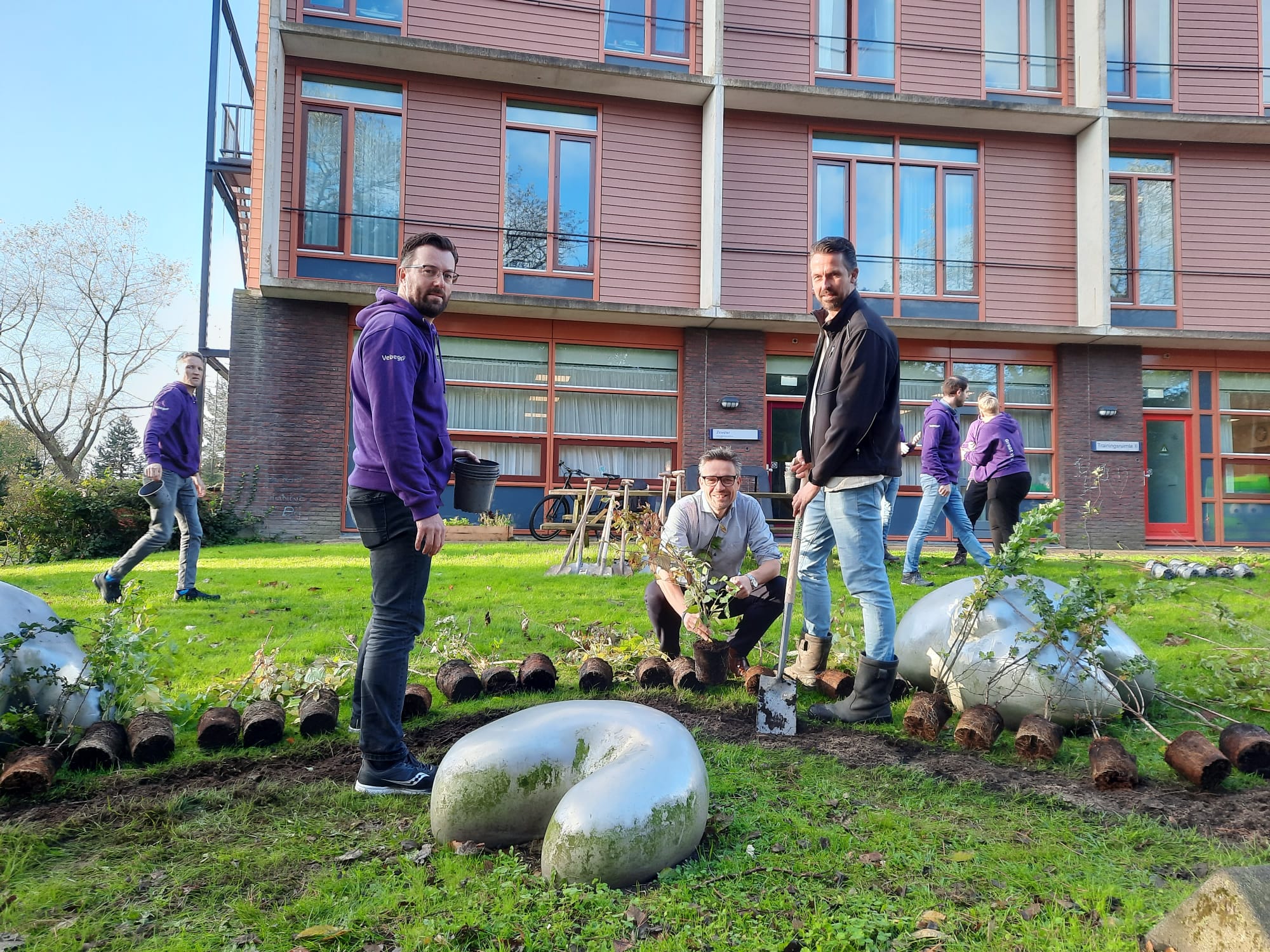 Mensen planten bomen in een groene ruimte voor een gebouw met rode kozijnen.