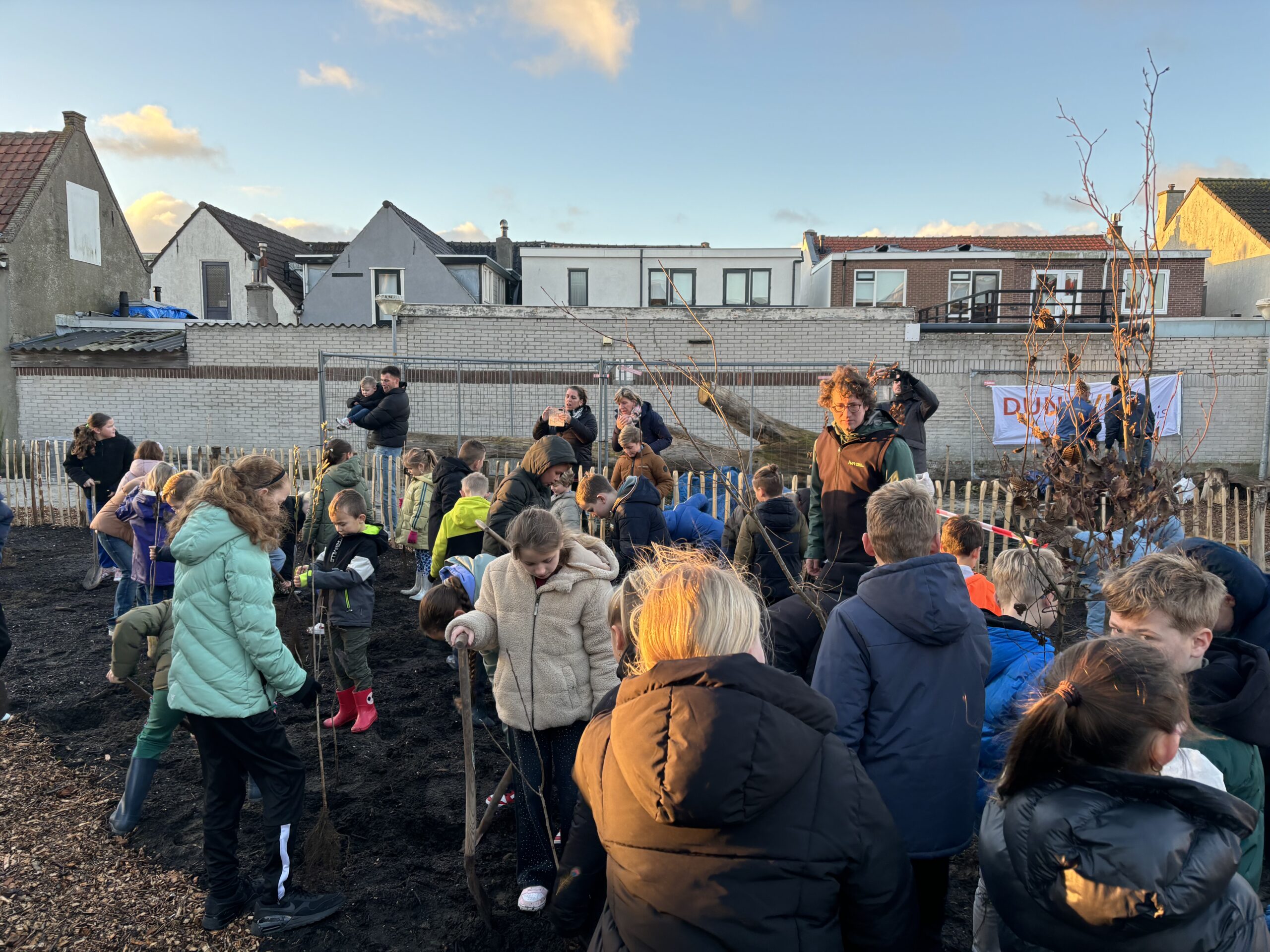 Groep kinderen plant bomen op een grasveld, begeleid door volwassenen, met huizen op de achtergrond.