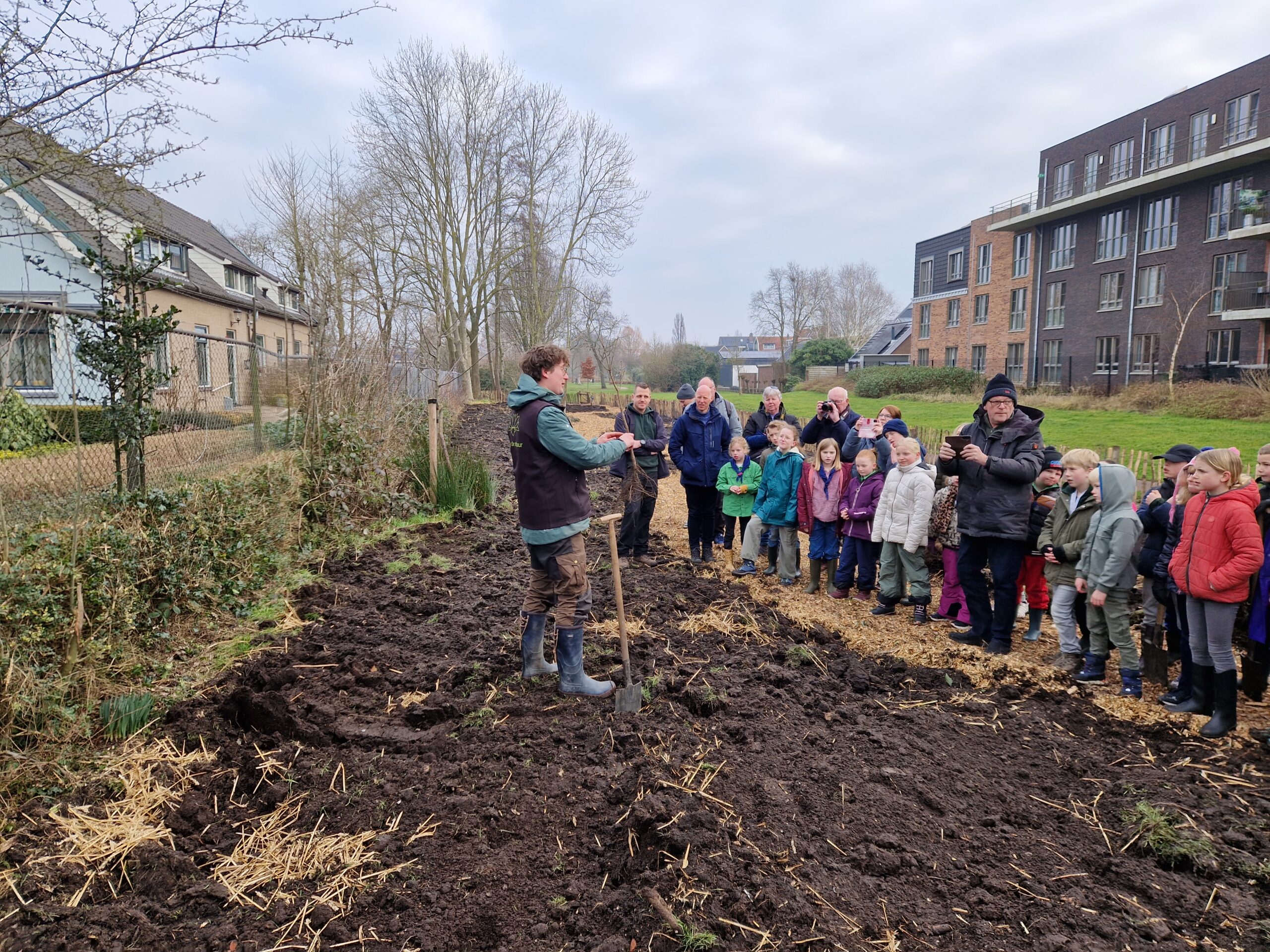 Man in regenlaarzen legt groep kinderen en volwassenen tuinieren uit in een moestuin naast gebouwen.