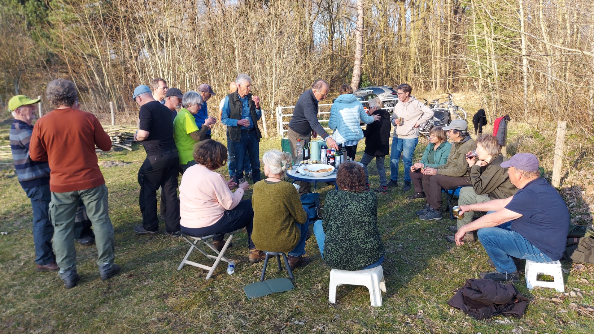 Groep mensen in een buitenbijeenkomst met drankjes en snacks op een grasveld bij een bosrand.