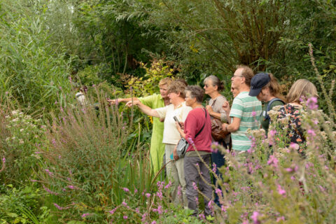 Een groep mensen observeert planten in een weelderige tuin, met bloemen en groen om hen heen.