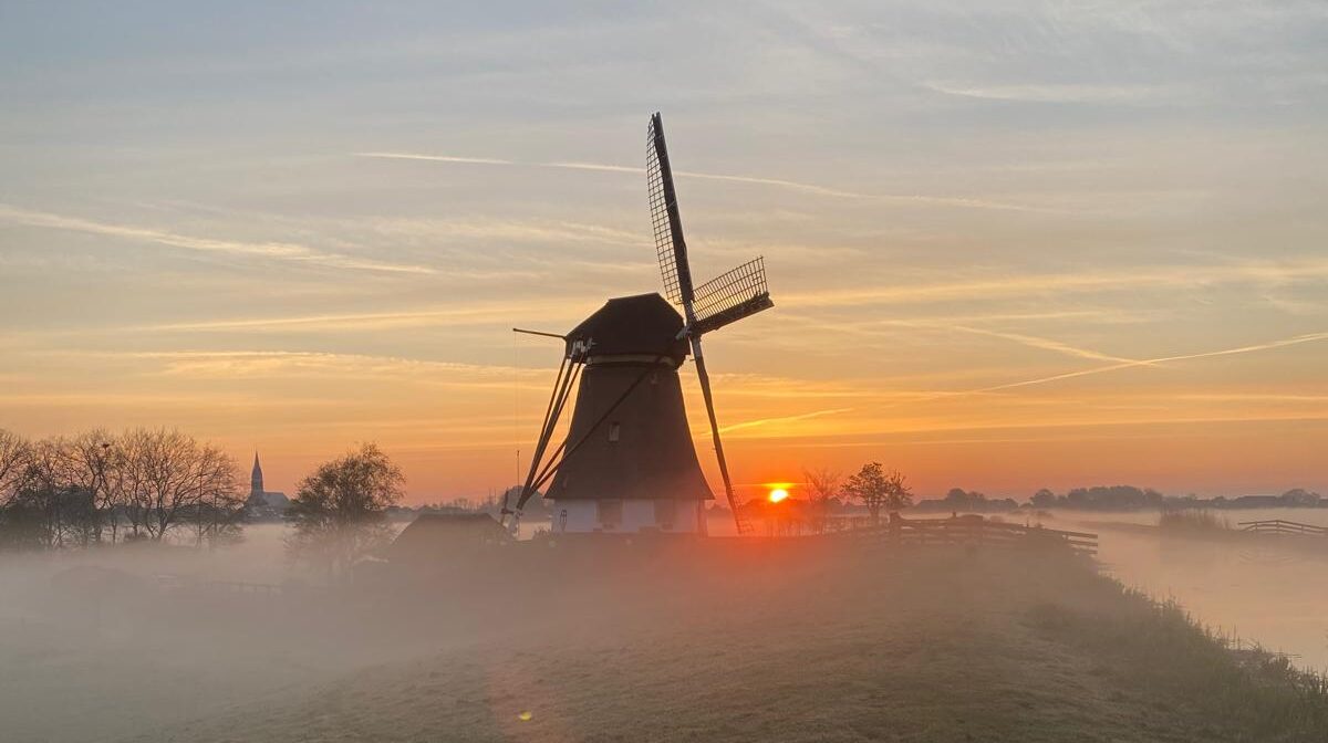 Windmolen bij zonsopgang, omgeven door mist, met bomen en kerktoren in de verte.