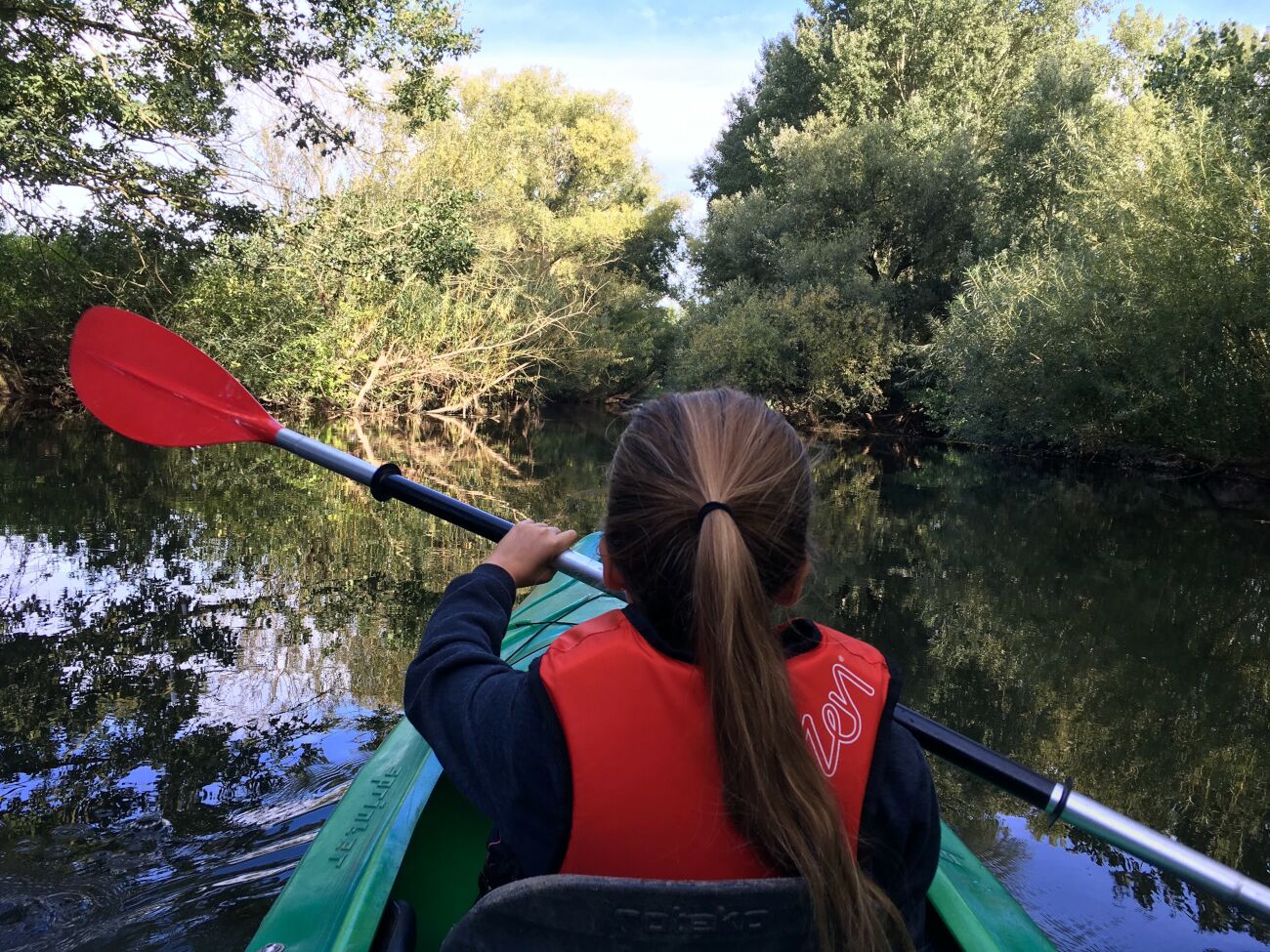 Persoon kajakt op smalle rivier omringd door weelderige groene bomen.
