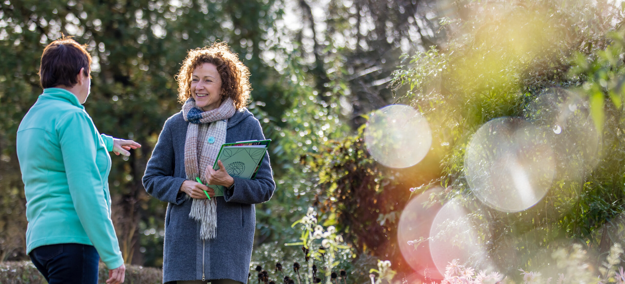Twee vrouwen in gesprek in een zonnige tuin, omringd door groen en bloemen.