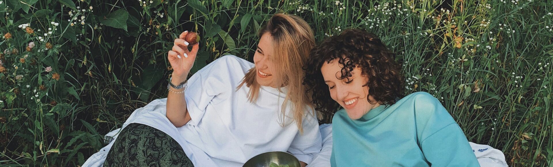 Twee personen zitten op een kleed in het gras, lachend met wat fruit.