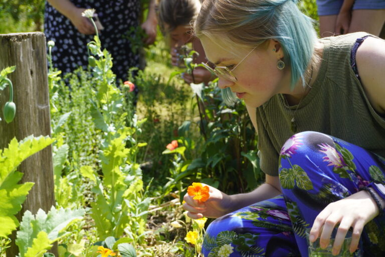 Persoon met blauw haar onderzoekt bloemen in een zonnige tuin met veel groen.