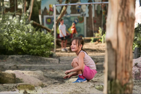 Meisje speelt lachend in de zandbak op een zonnige dag; kinderen spelen op de achtergrond.