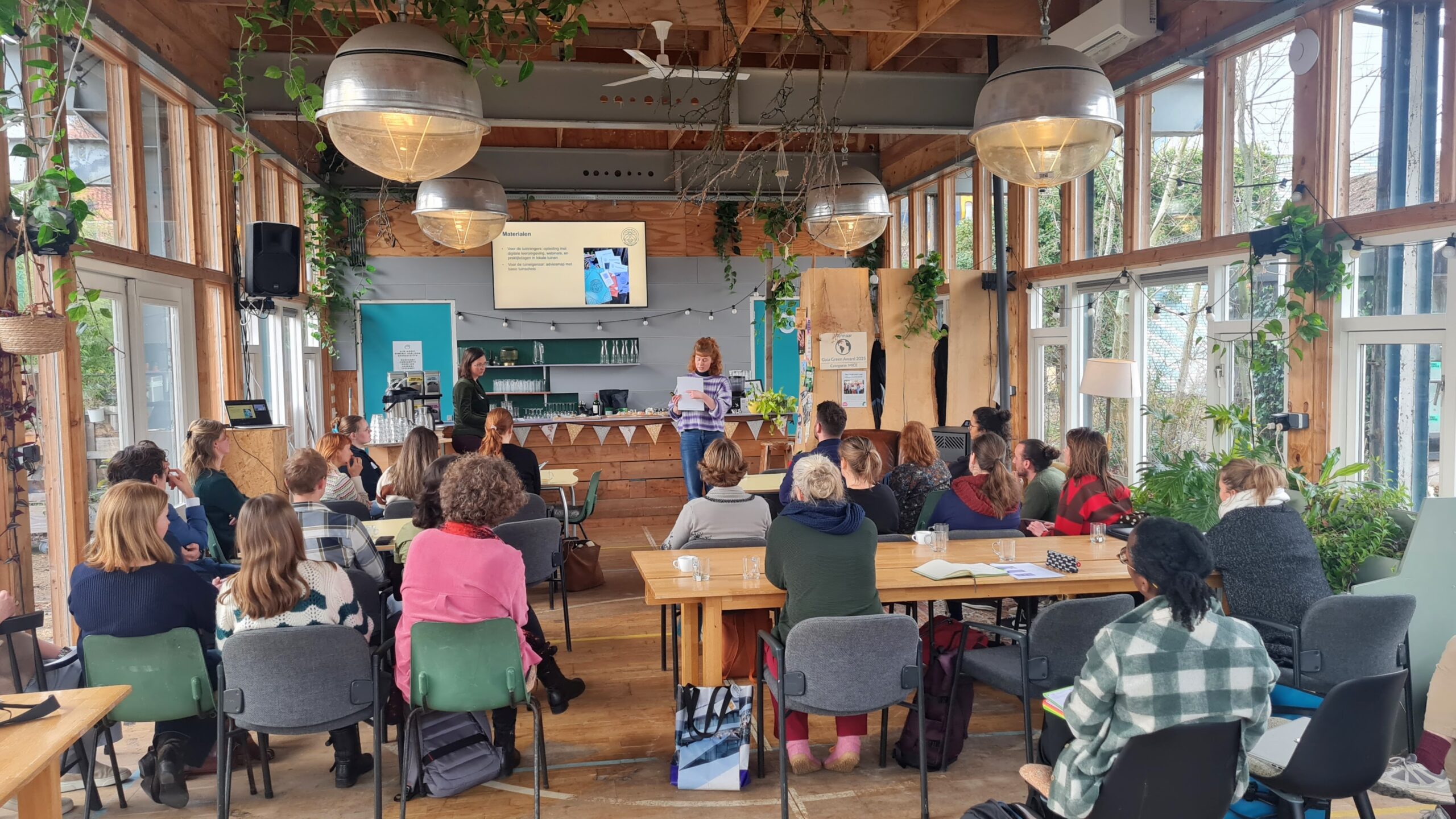 Een presentatie in een lichte ruimte met planten; publiek kijkt naar spreker en scherm.