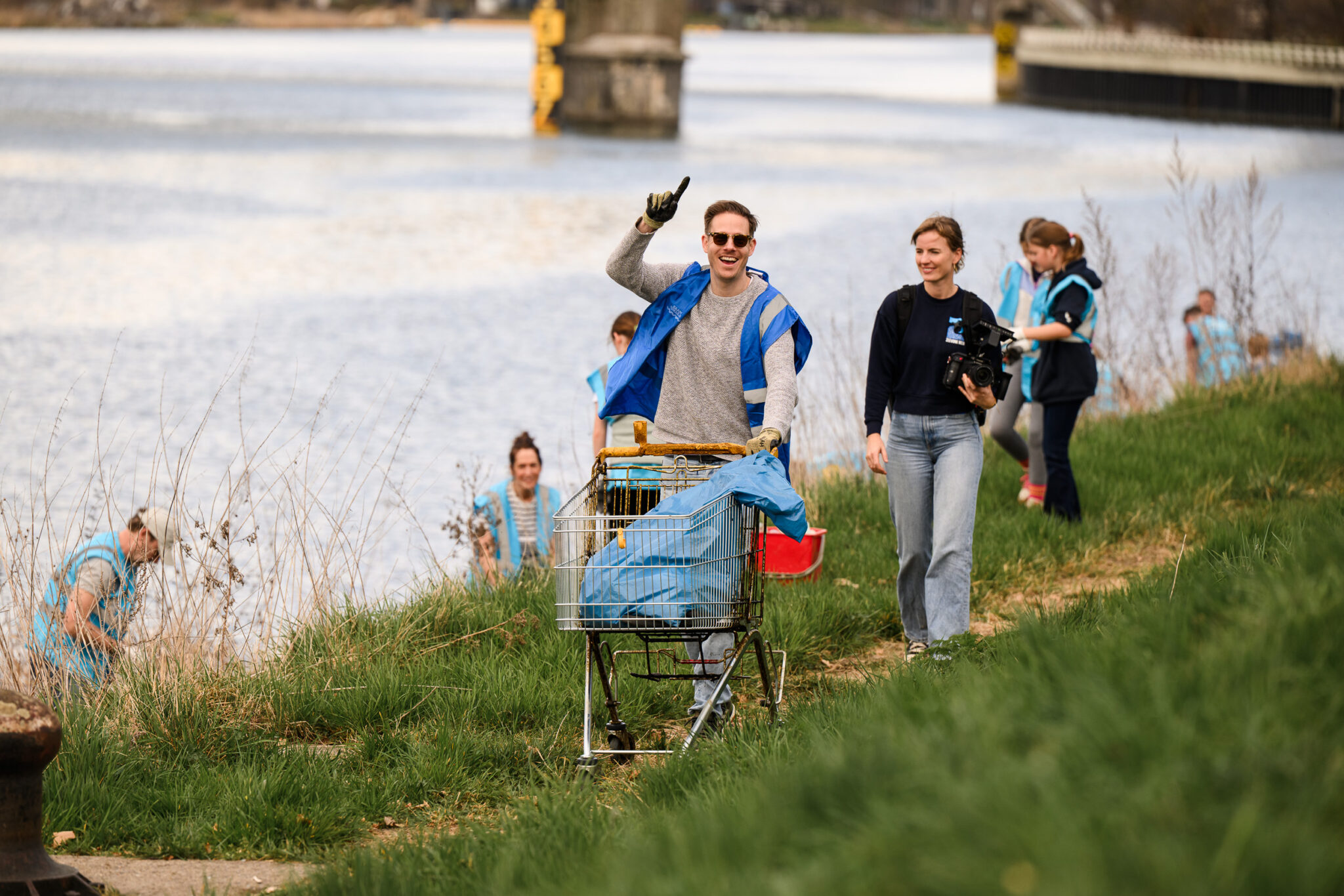 Mensen in blauwe hesjes ruimen afval op langs een rivier; een man duwt een winkelwagentje vol vuilnis.