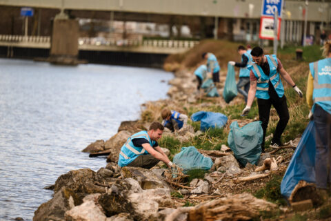 Mensen in blauwe hesjes ruimen afval op langs een rivier onder een brug.