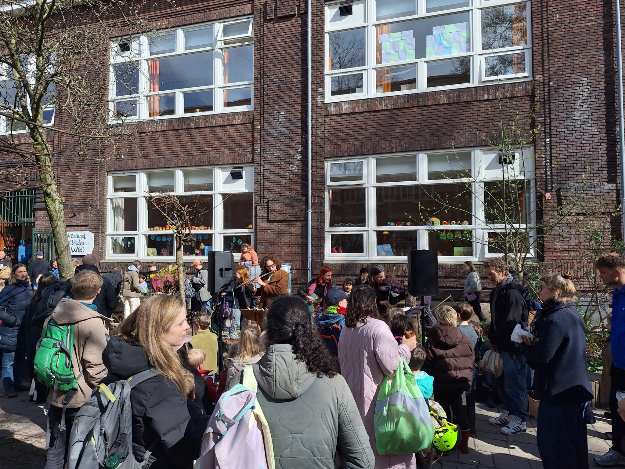 Mensen verzamelen zich buiten een school voor een optreden of evenement op een zonnige dag.