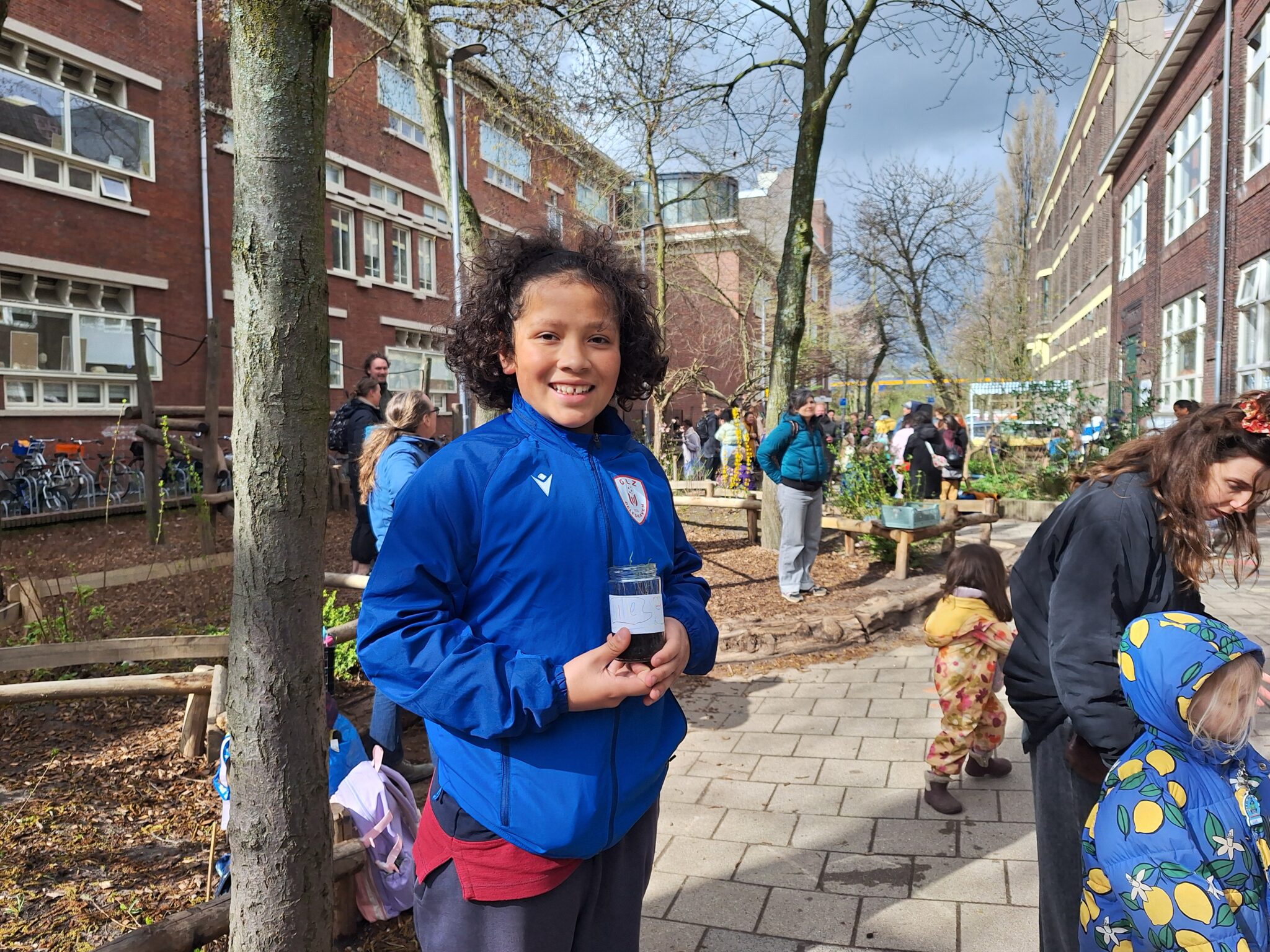Jongen in blauwe jas glimlacht en houdt een klein potje vast. Mensen staan buiten bij een bakfiets.