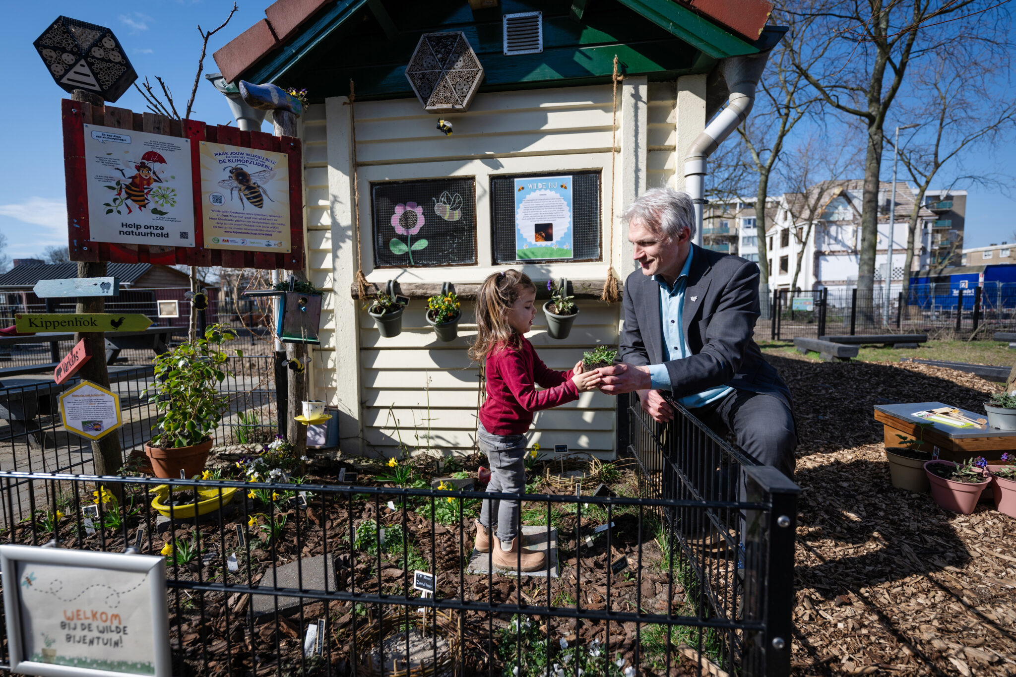 Man en kind tuinieren bij een klein gebouwtje in een zonnige speeltuin.