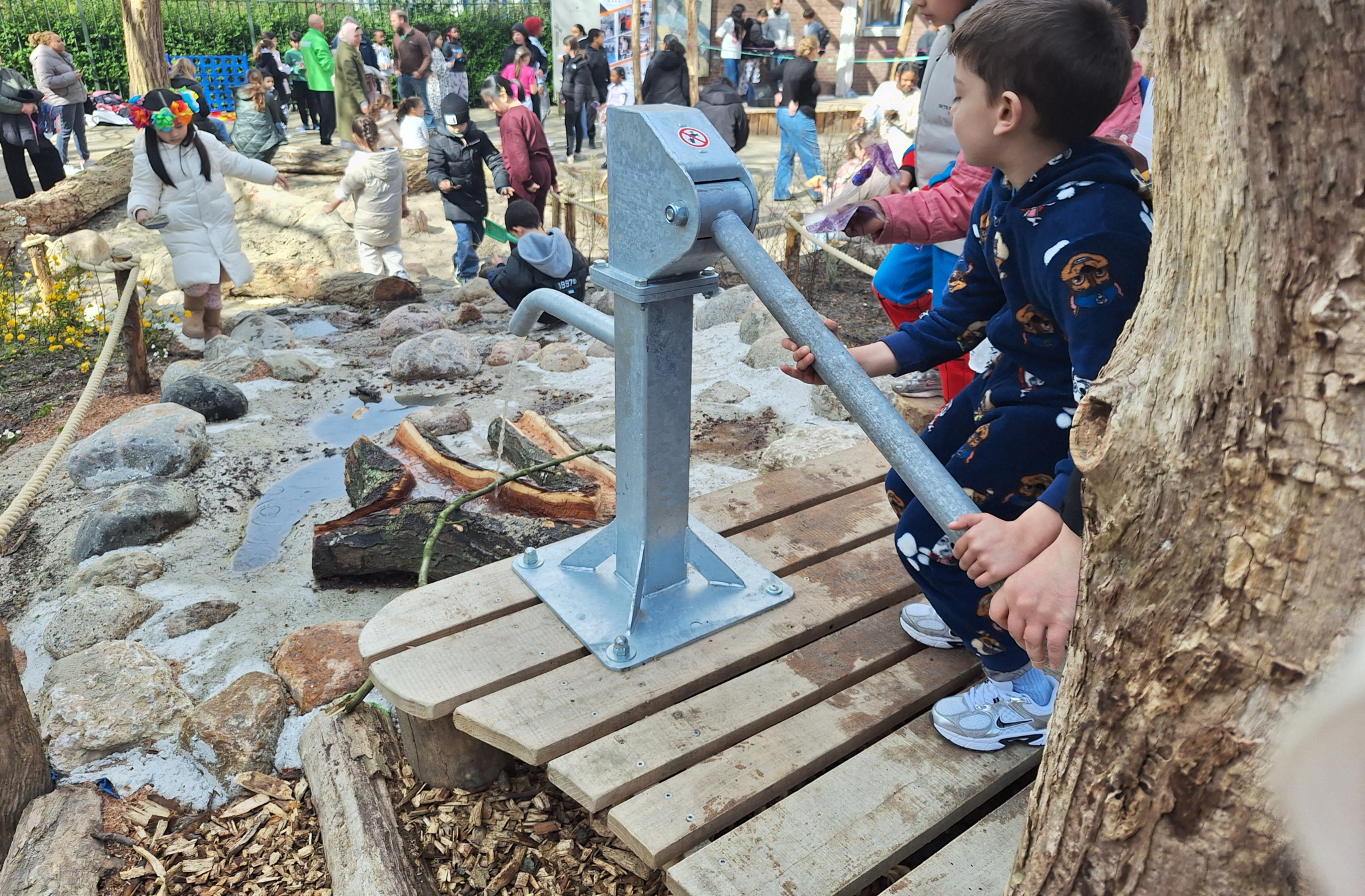 Kinderen spelen bij een waterpomp in een speeltuin, omringd door natuur en volwassenen.