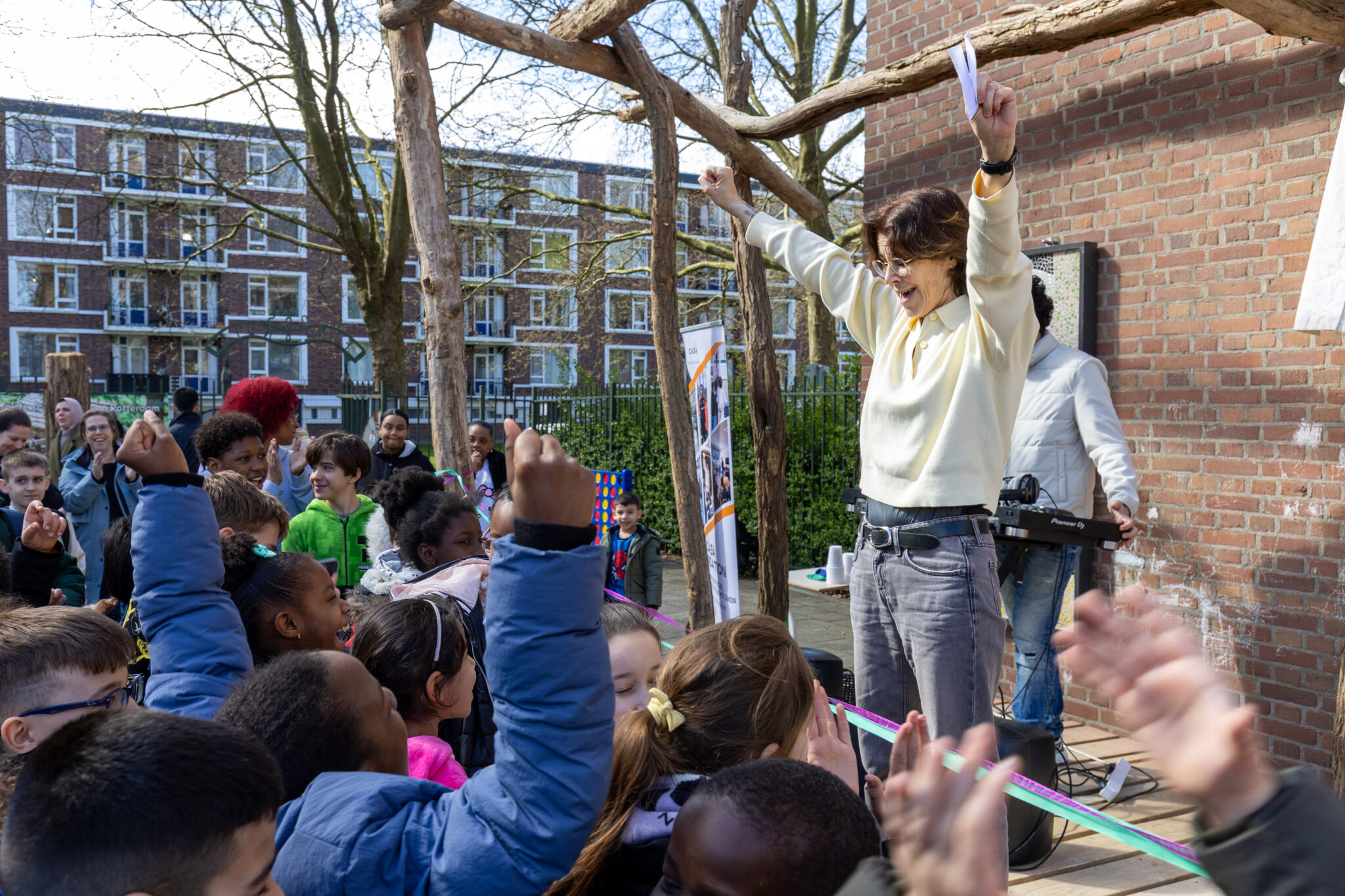 Een vrouw juicht met opgeheven armen voor een groep enthousiaste kinderen buiten.