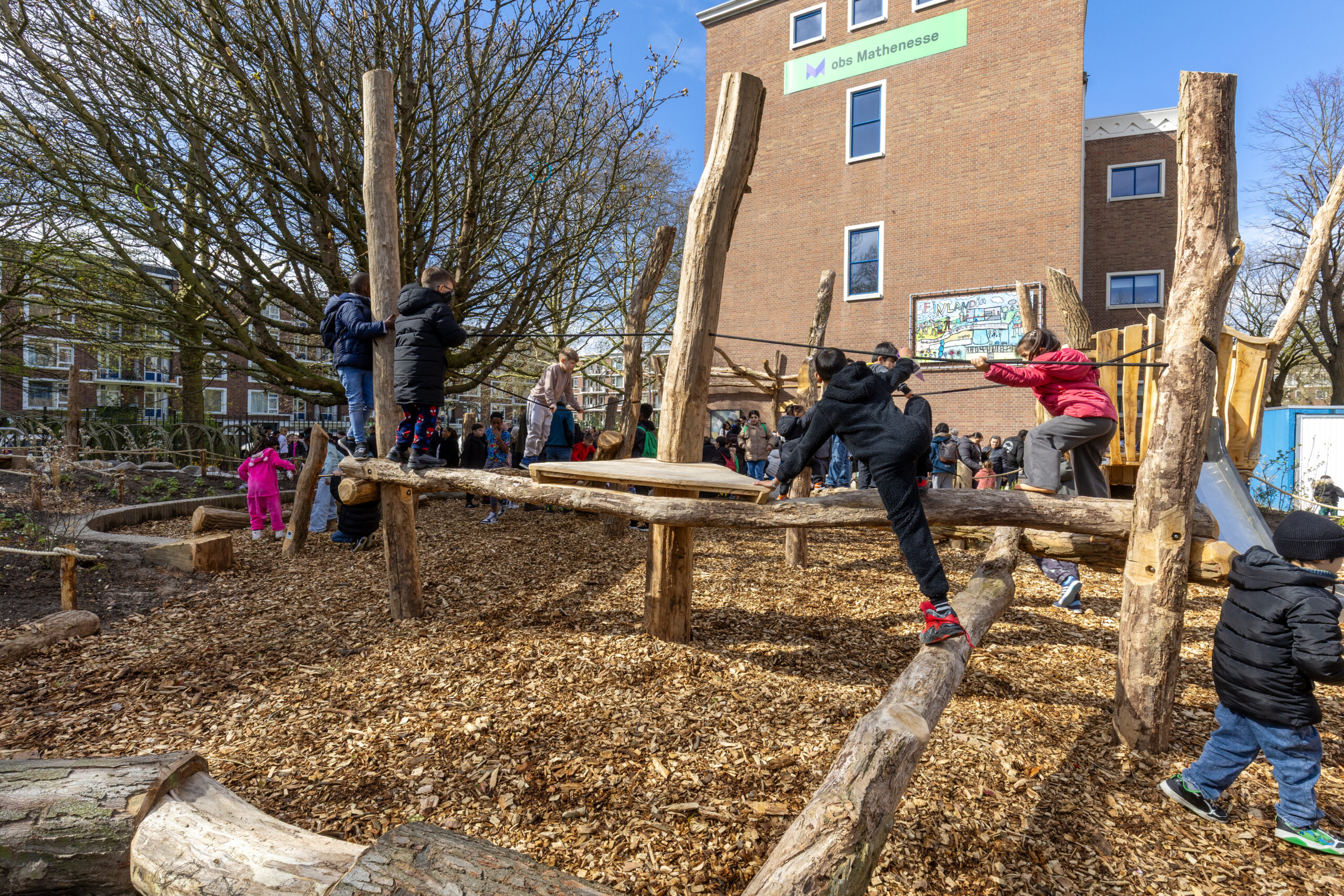 Kinderen spelen op houten klimtoestellen op een schoolplein met houtsnippers en een boom.